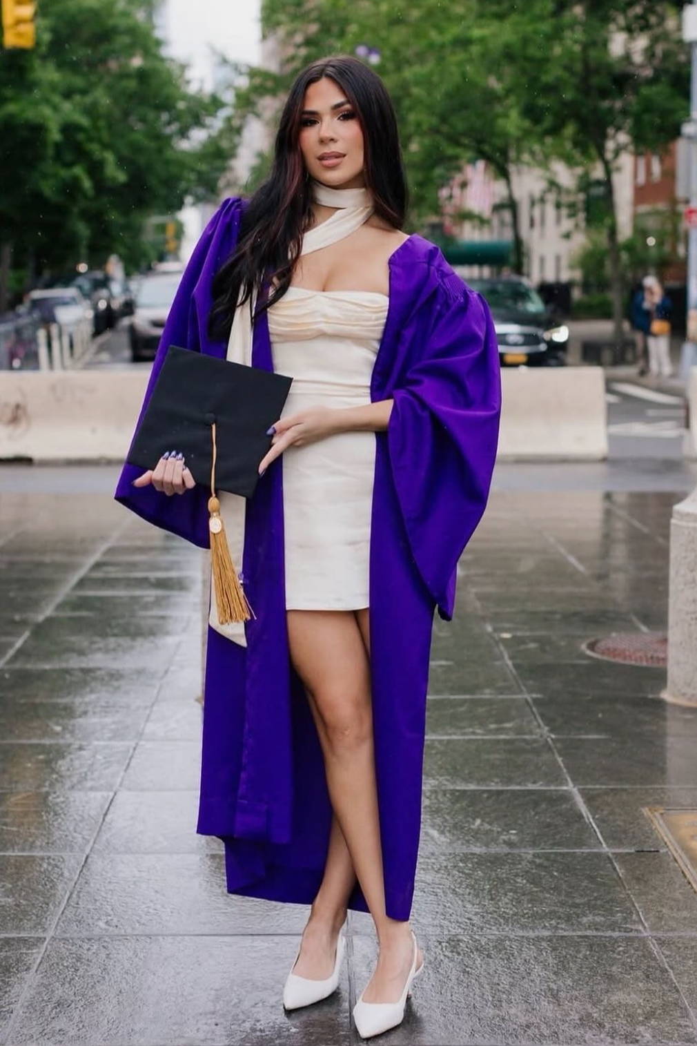 Young woman in graduation gown holding a diploma, standing on a city street.