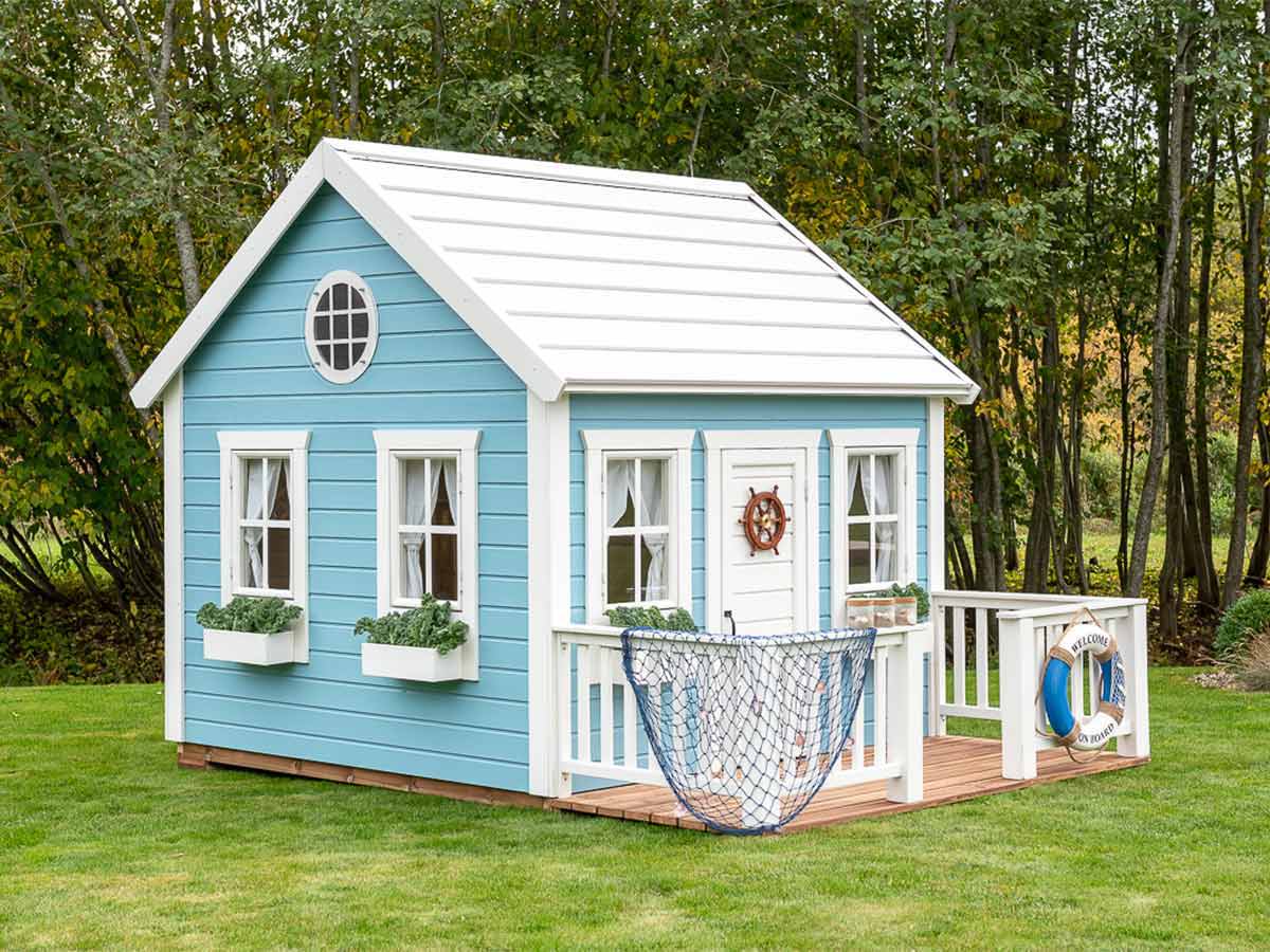 Wooden Playhouse in blue color with a white roof and a porch and flower boxes on green lawn with forest on the background by WholeWoodPlayhouses