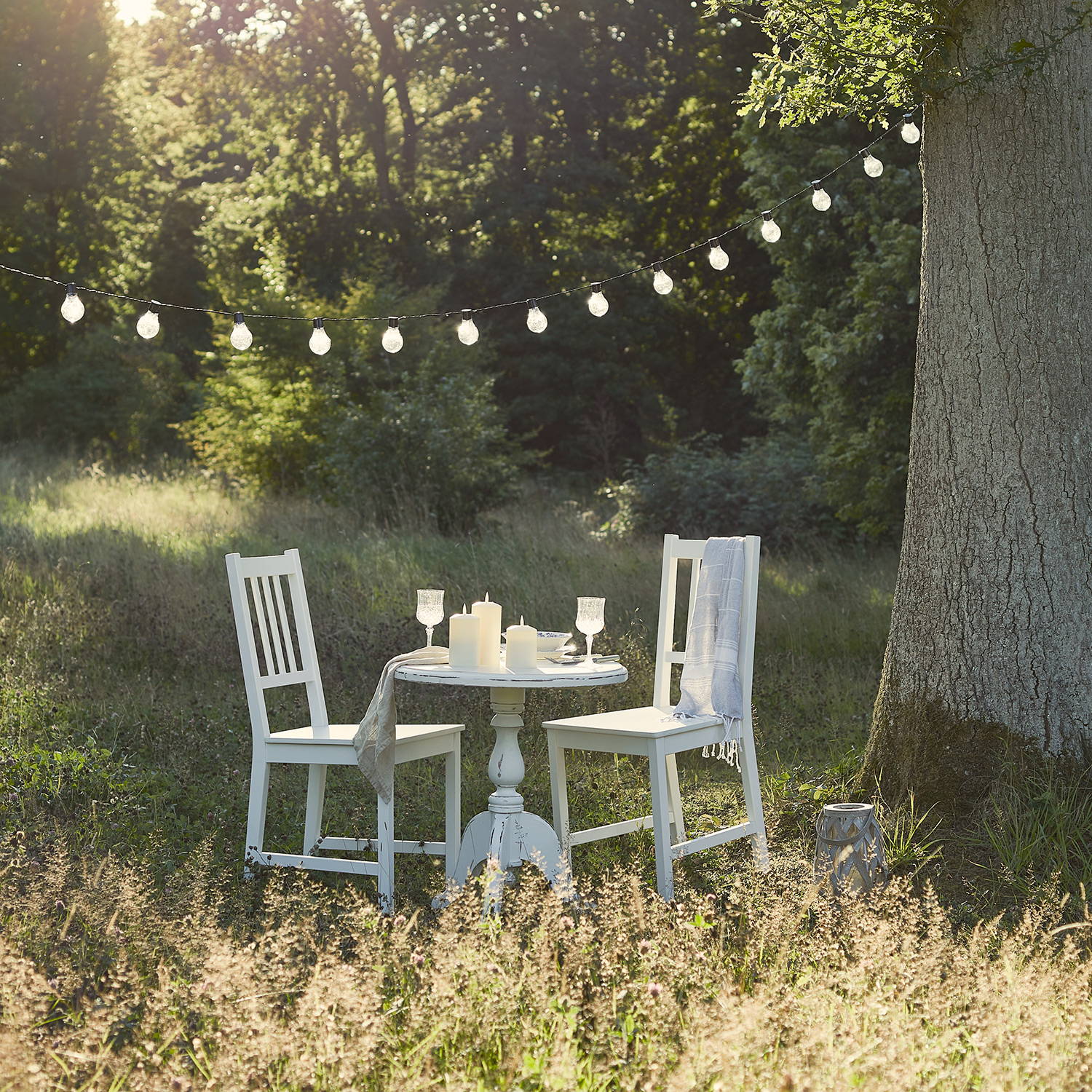 An alfresco dining set up in a meadow with festoon lights, candles and drinks.