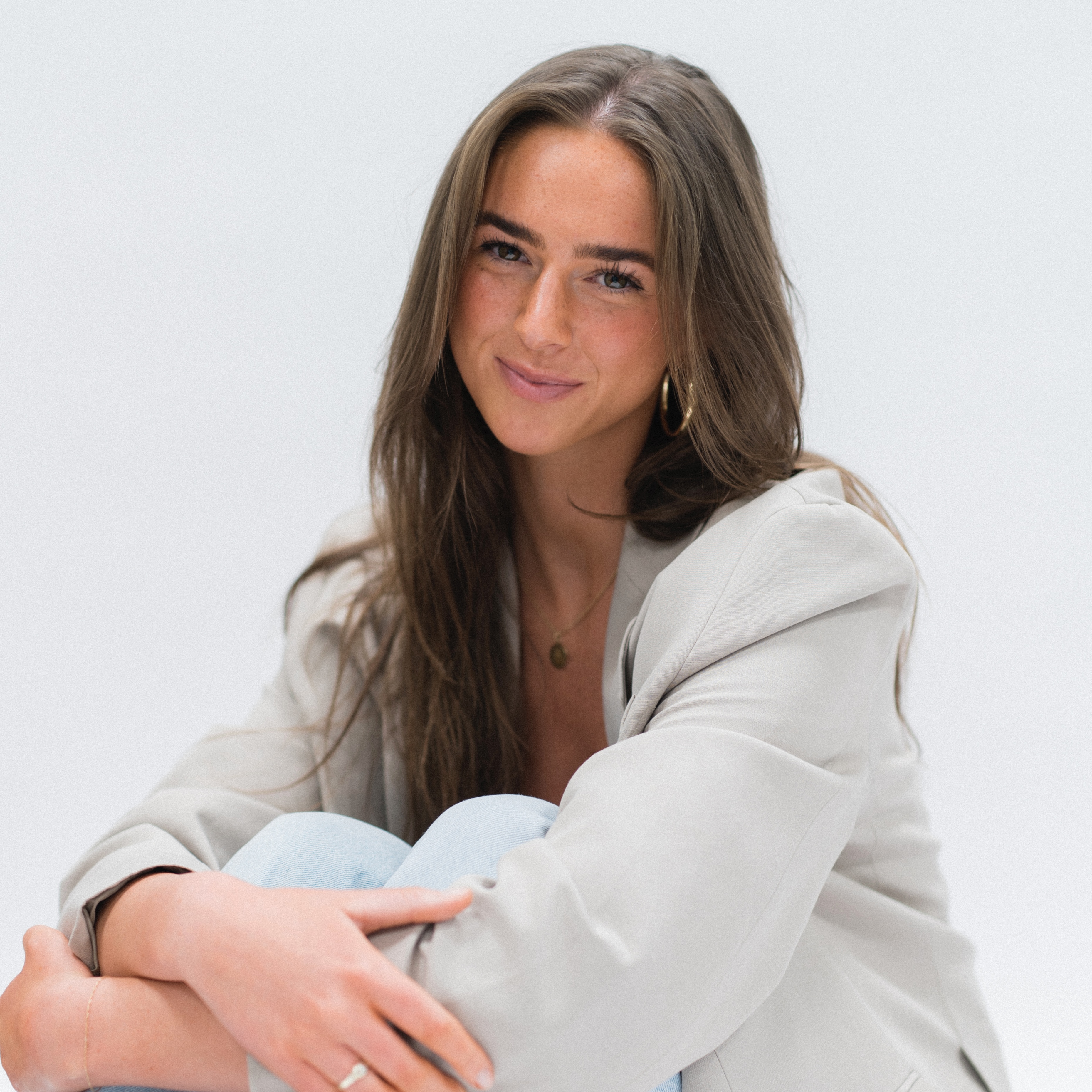Young woman smiling, sitting with knees drawn up, wearing a light blazer.