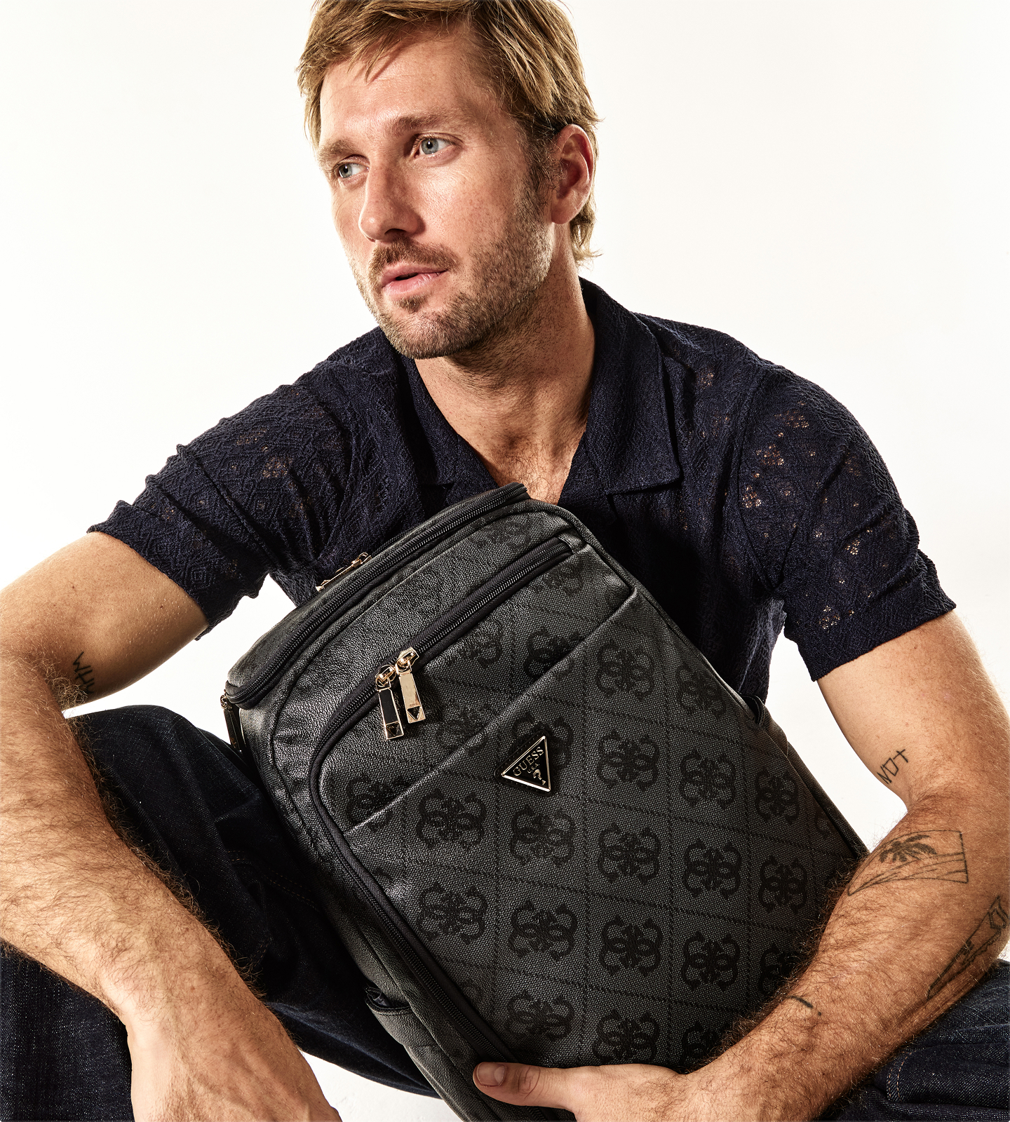 Man in dark shirt holding a GUESS backpack, seated against a light background.