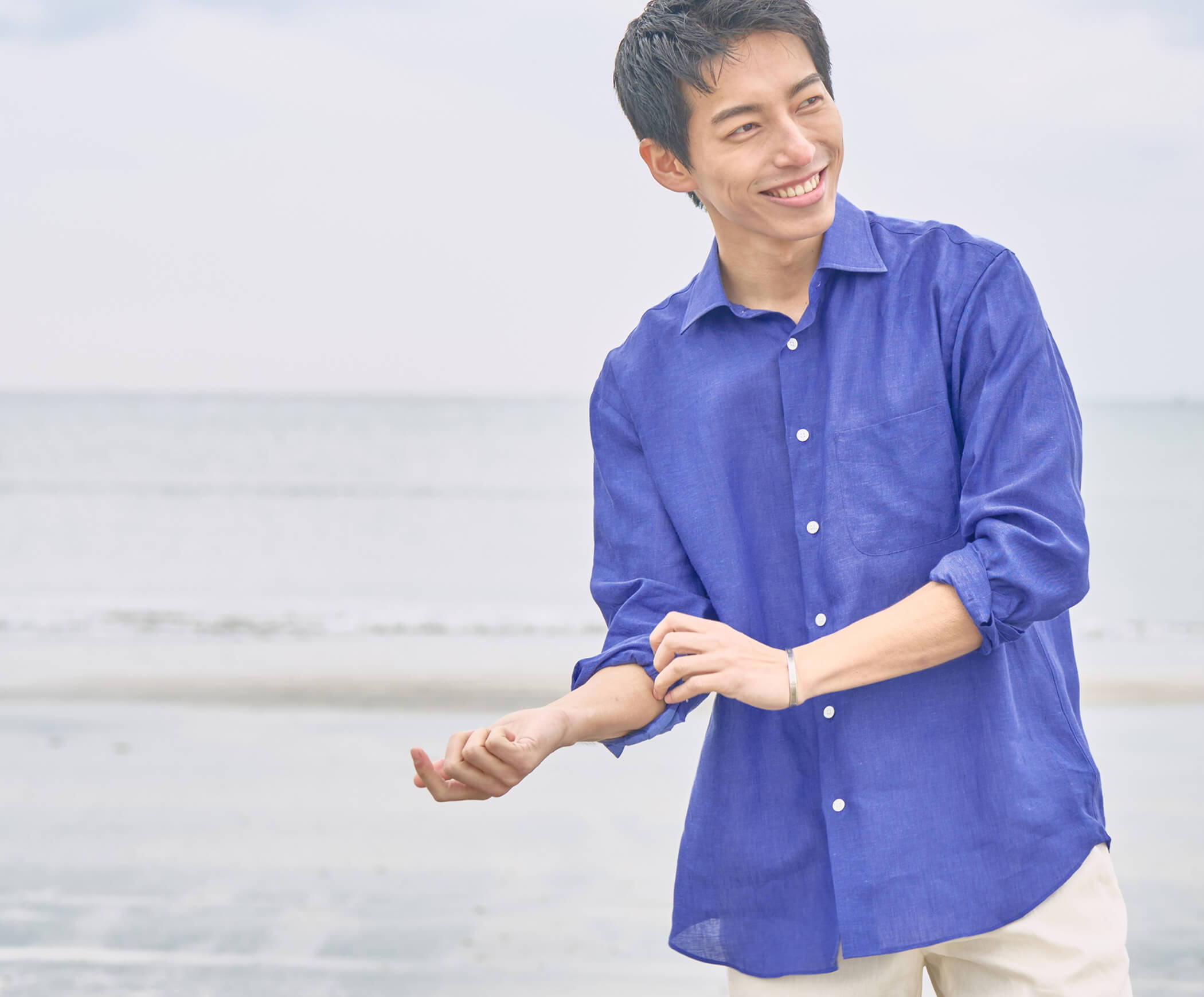 A young man in a blue shirt smiles by the beach.