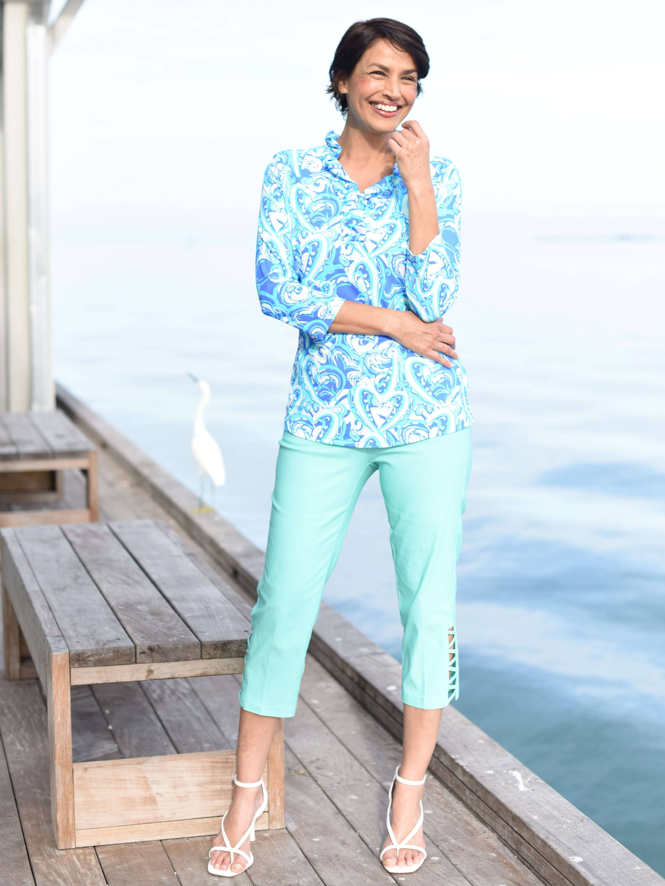 A woman with short dark hair wears a blue printed top and seafoam colored pants as she stands on a dock with a heron in the background