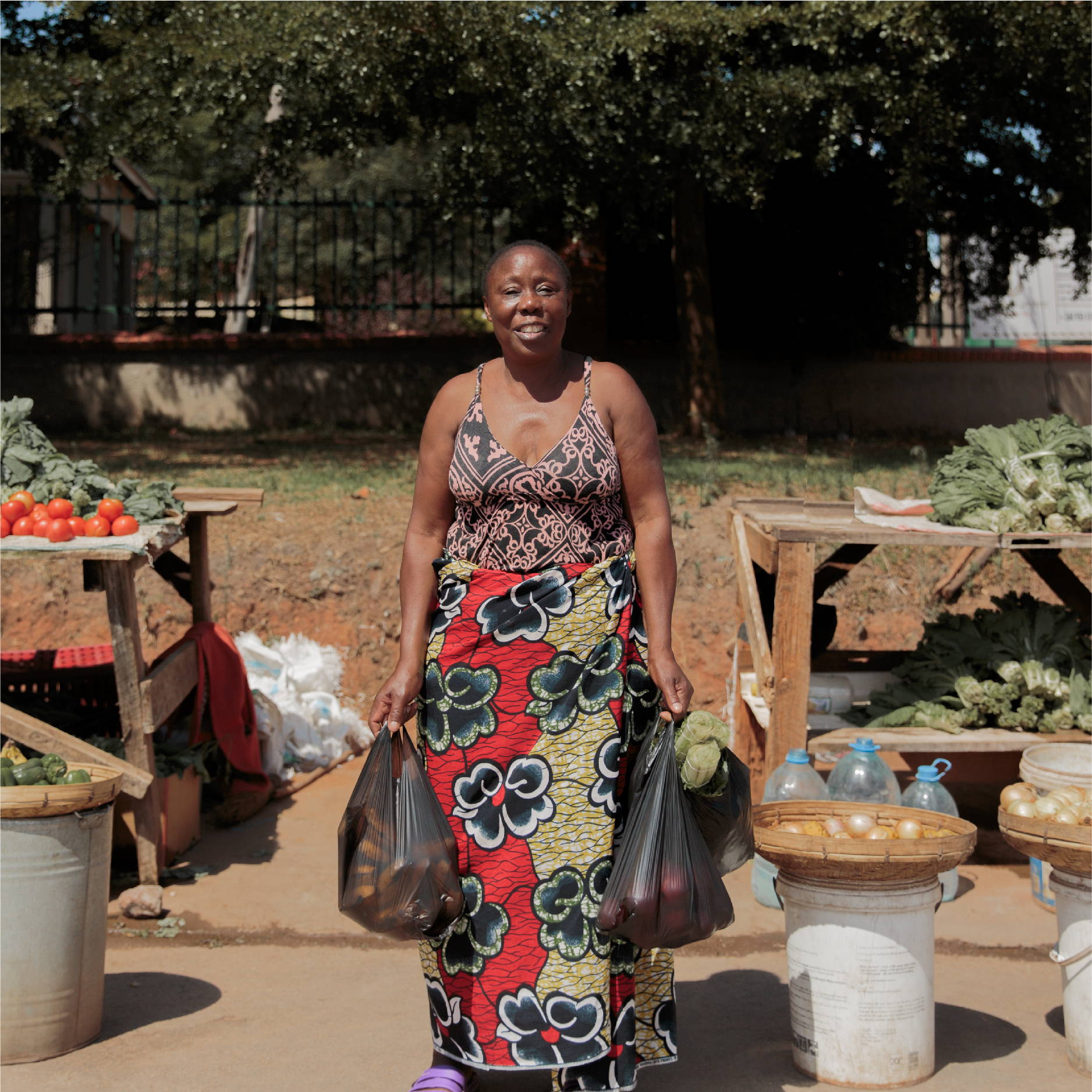 A woman stands smiling in front of two street vendors selling fresh local produce in Zambia. She holds two full black bags. 