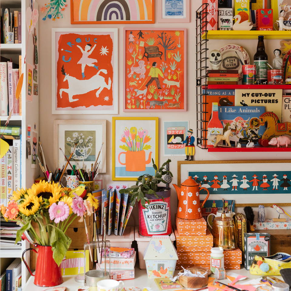 A desk filled with colourful flowers, stationary, trinkets and books with colourful framed Printed Peanut pictures on the wall behind.