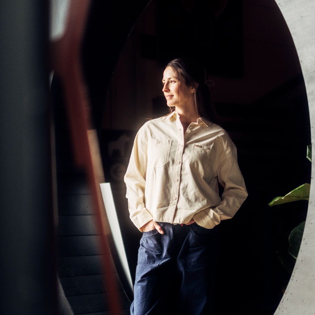 A person in a light shirt stands near a mirror, looking thoughtfully.