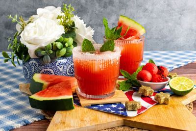 Two cocktails with watermelon, surrounded by fruit and flowers on a table.