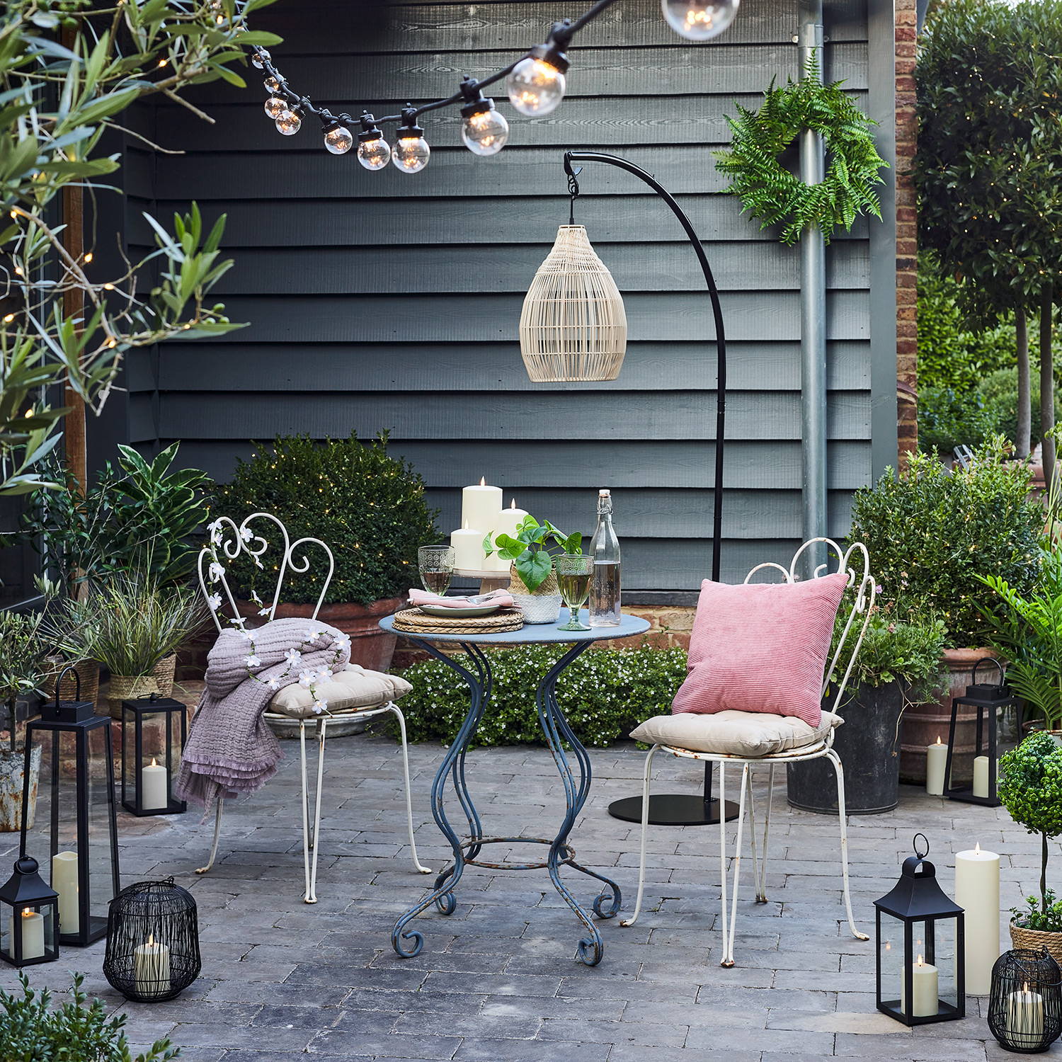 An outdoor patio dining area with lanterns, festoon lights, and a rattan hanging pendant.