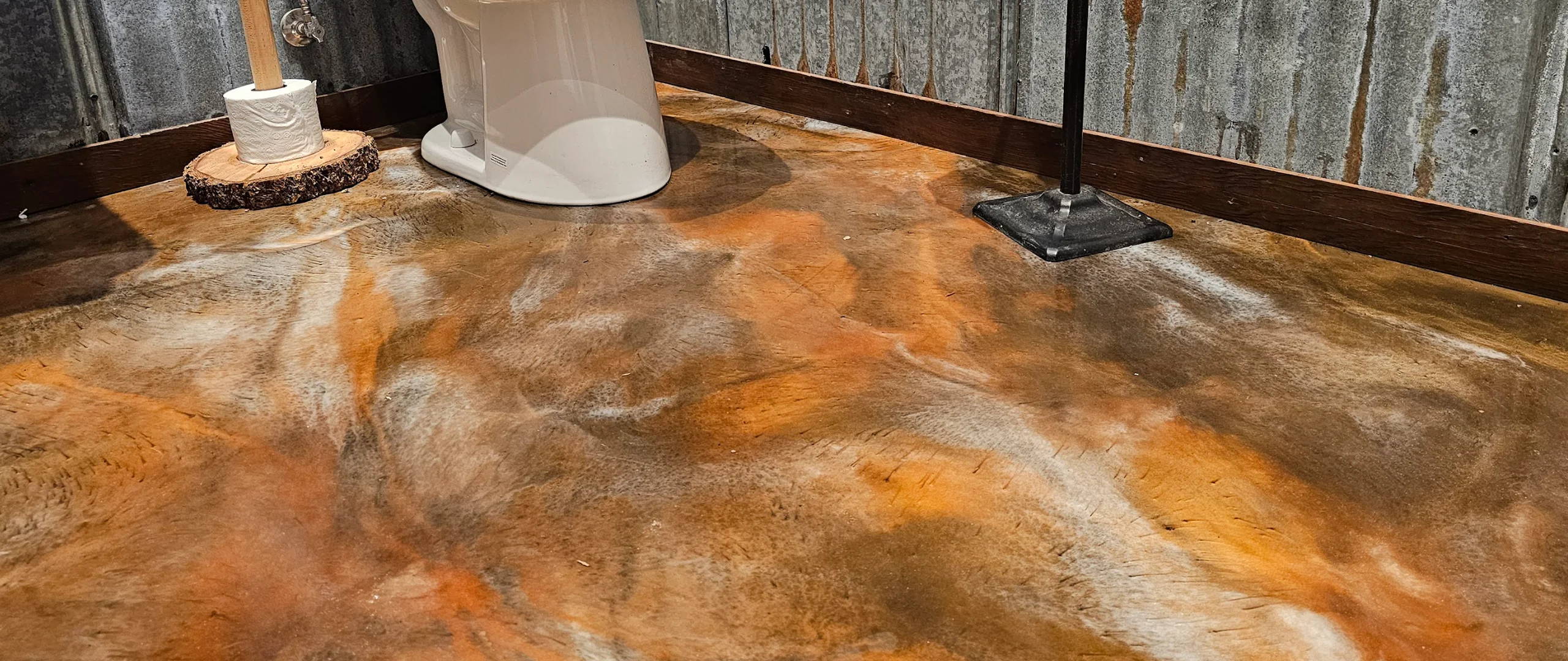 Unique bathroom with concrete floor, orange-brown stain, white pedestal sink, black broom, corrugated metal walls, wooden baseboards. 