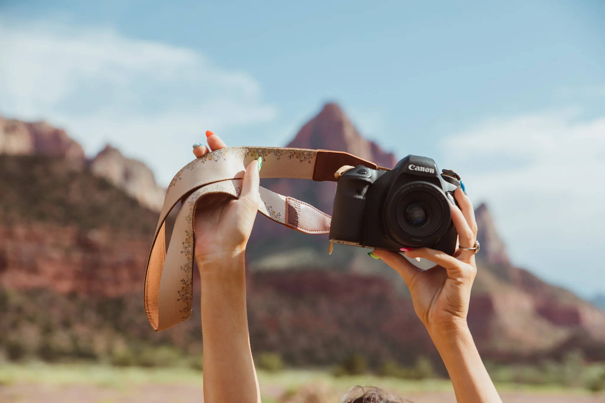 woman holding up a camera with a flower camera strap in front of mountain background and blue sky.