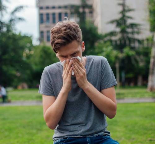 Ein junger Mann sitzt im Park und schnäuzt sich seine Nase, die aufgrund seines Heuschnupfens läuft oder verstopft 