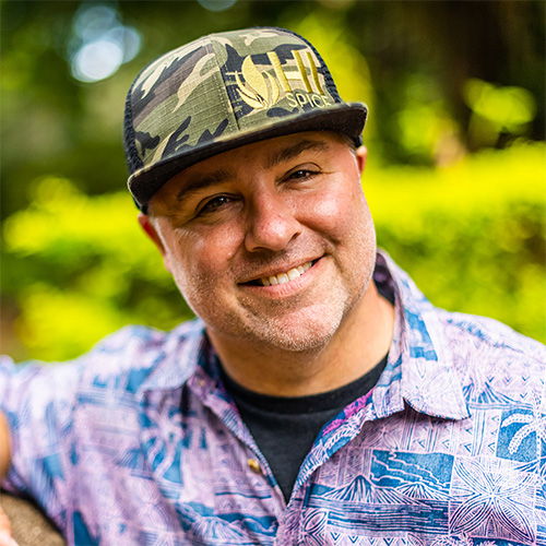 Smiling man in a floral shirt and camo hat outdoors.