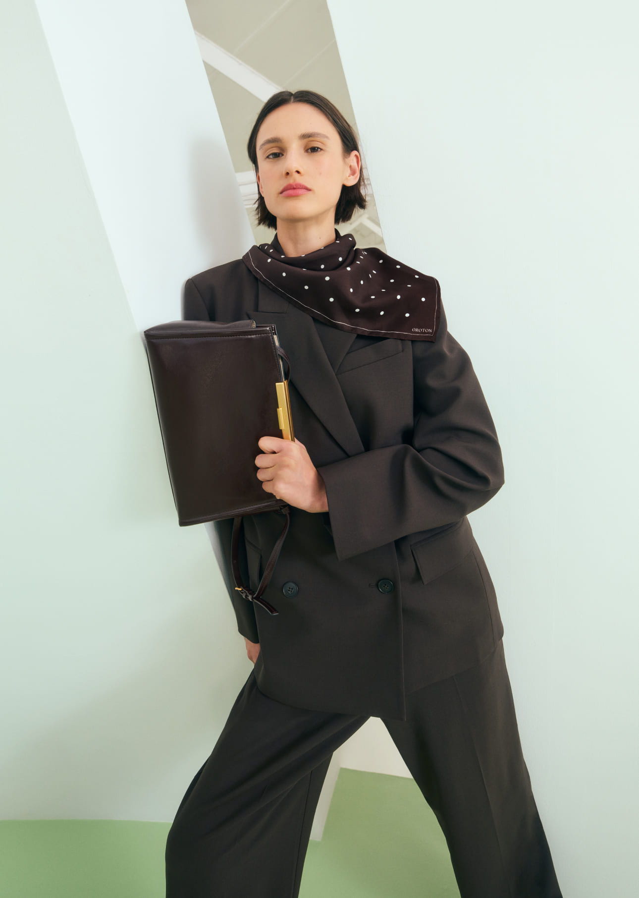 Woman in stylish outfit holding a clutch, standing against a pastel background.