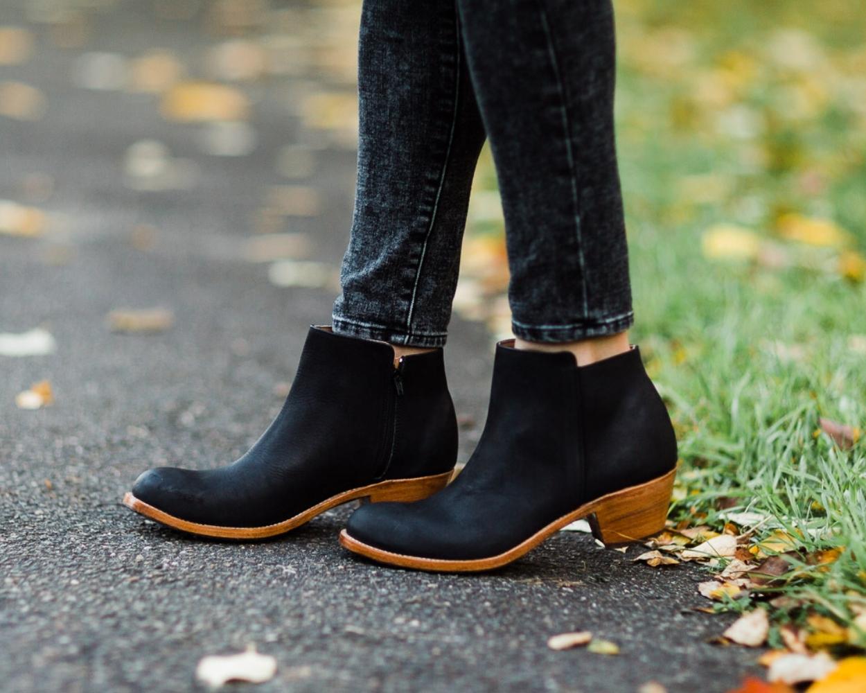 An outdoor lifestyle shot from the calves down of a person standing on an asphalt path scattered with autumn leaves. They are wearing dark grey or black slim-fit denim jeans and a pair of classic black leather ankle boots featuring a subtle side zipper, a rounded toe, and a contrasting light tan low stacked leather heel.