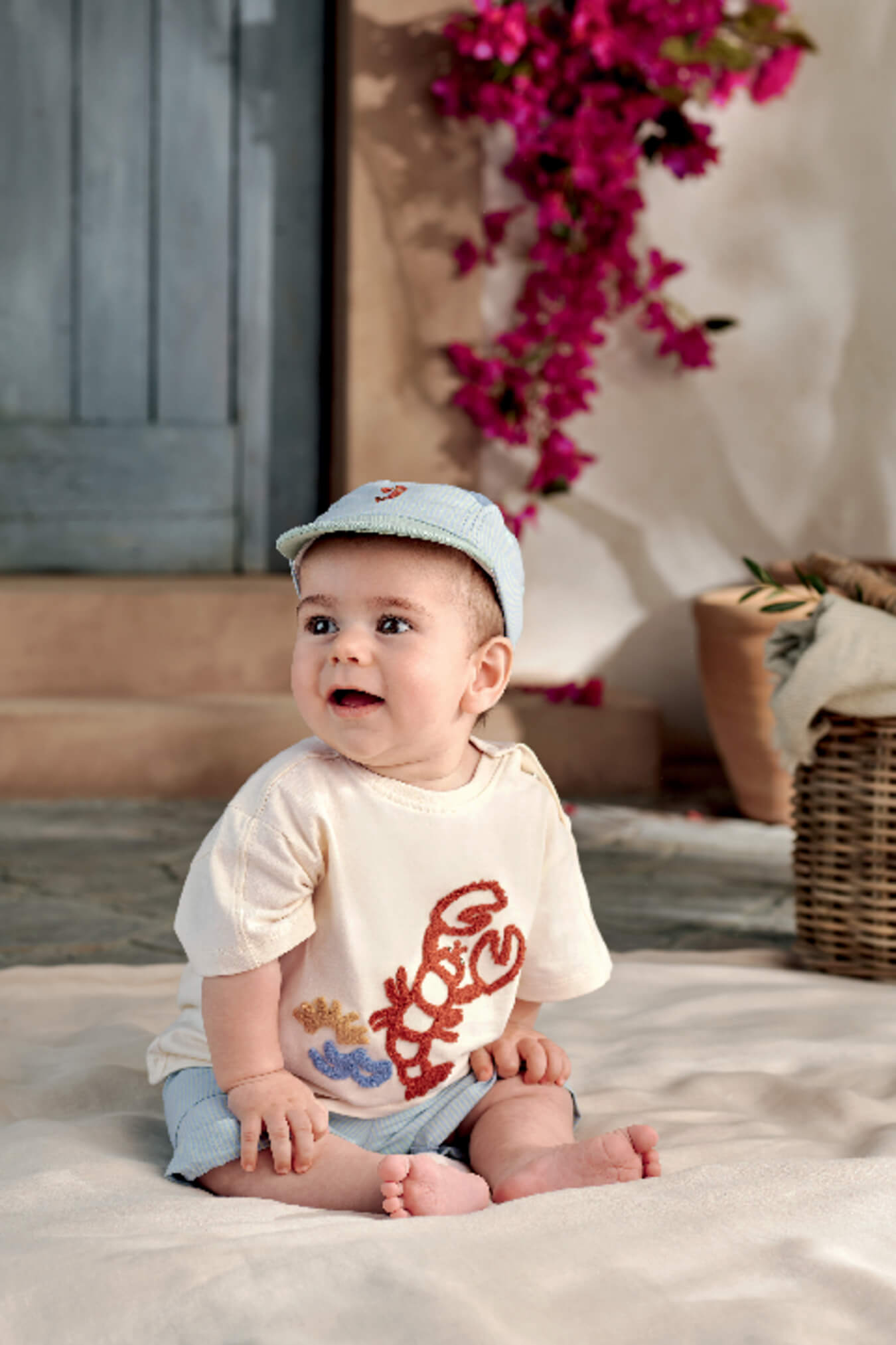 Baby sitting on the floor, wearing a shirt and cap, surrounded by flowers.