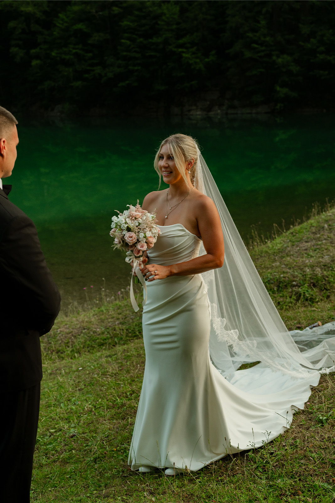 Bride in white dress holding bouquet, standing by water, looking at groom.