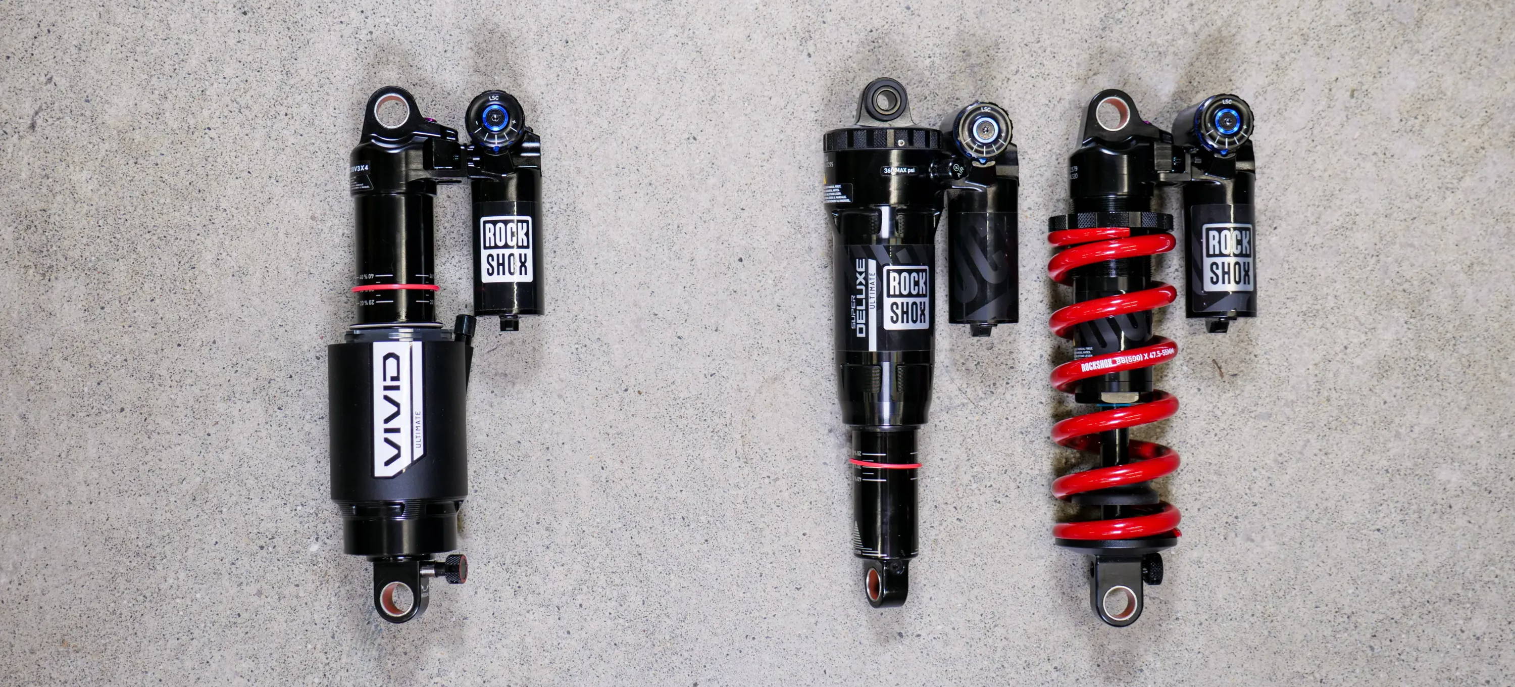a comparison of rockshox super deluxe ultimate and super deluxe coil ultimate and vivid rear mountain bike shocks laying on concrete 
