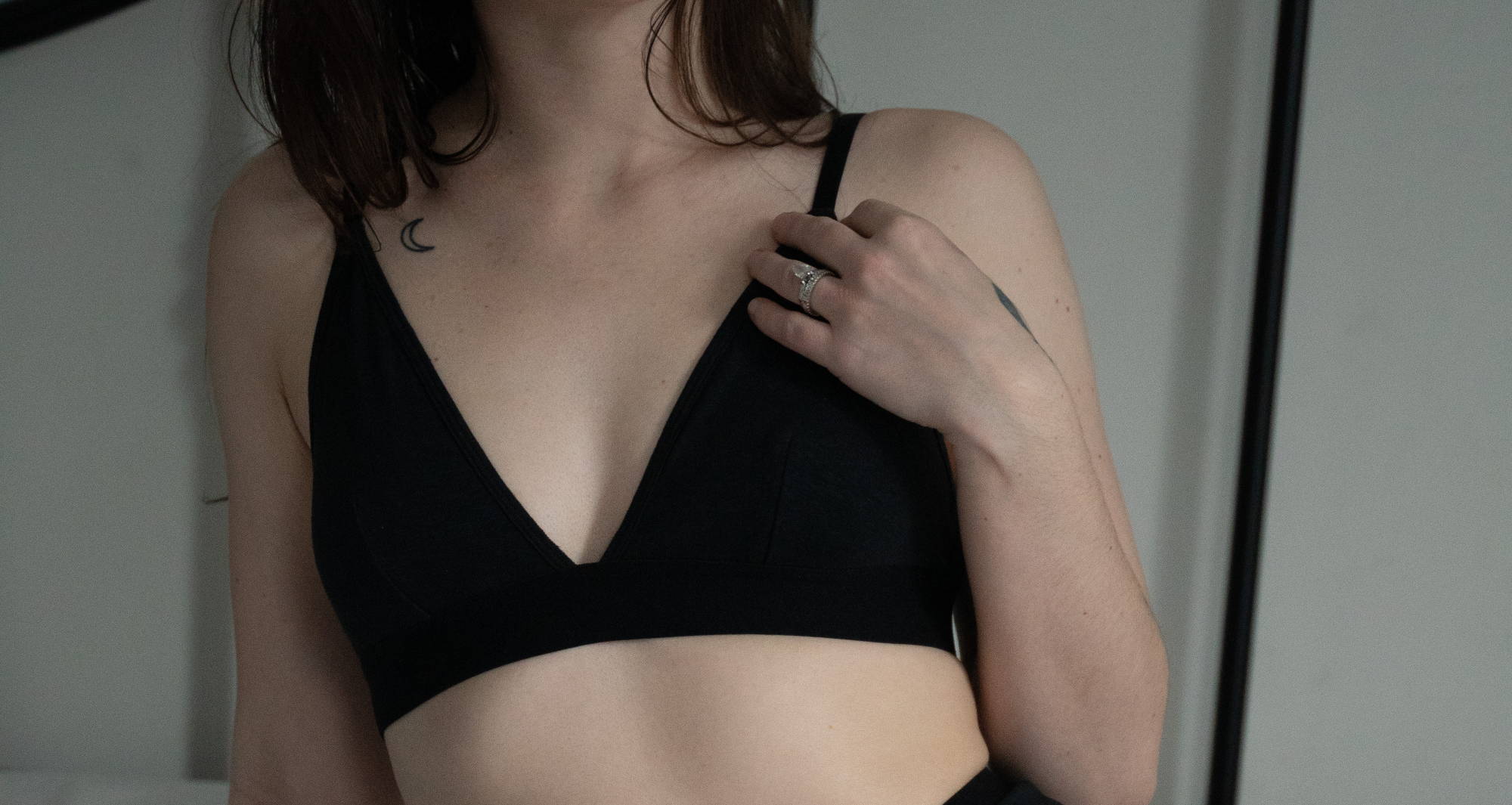 woman adjusts black bralette and leans against a white wall 