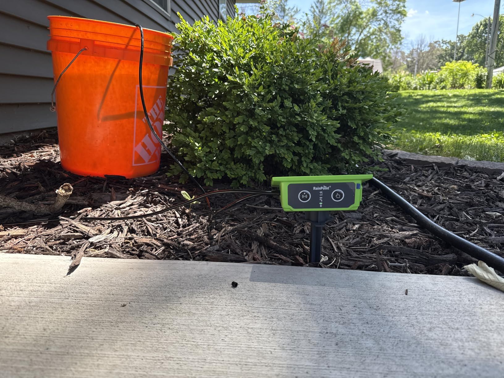 An orange bucket next to a bush and a green device on the ground.