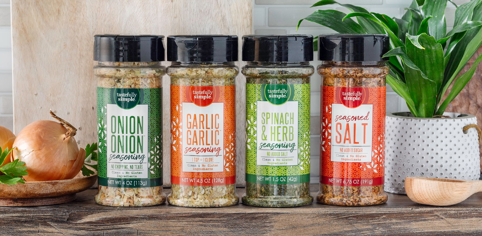 Four Tastefully Simple seasoning bottles—Onion Onion™, Garlic Garlic™, Spinach & Herb, and Seasoned Salt—displayed on a kitchen counter.