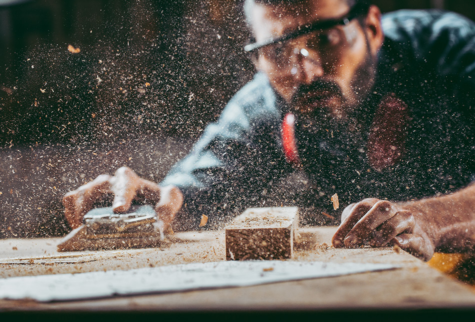 A carpenter sanding wood, surrounded by dust and wood shavings.