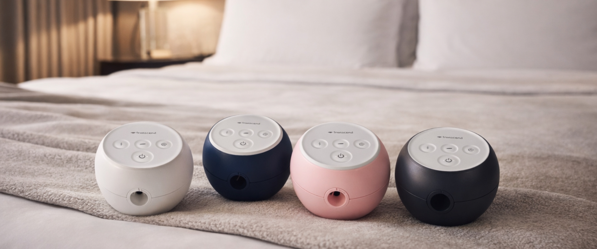 Four sound machines in white, navy, pink, and black on a bed.
