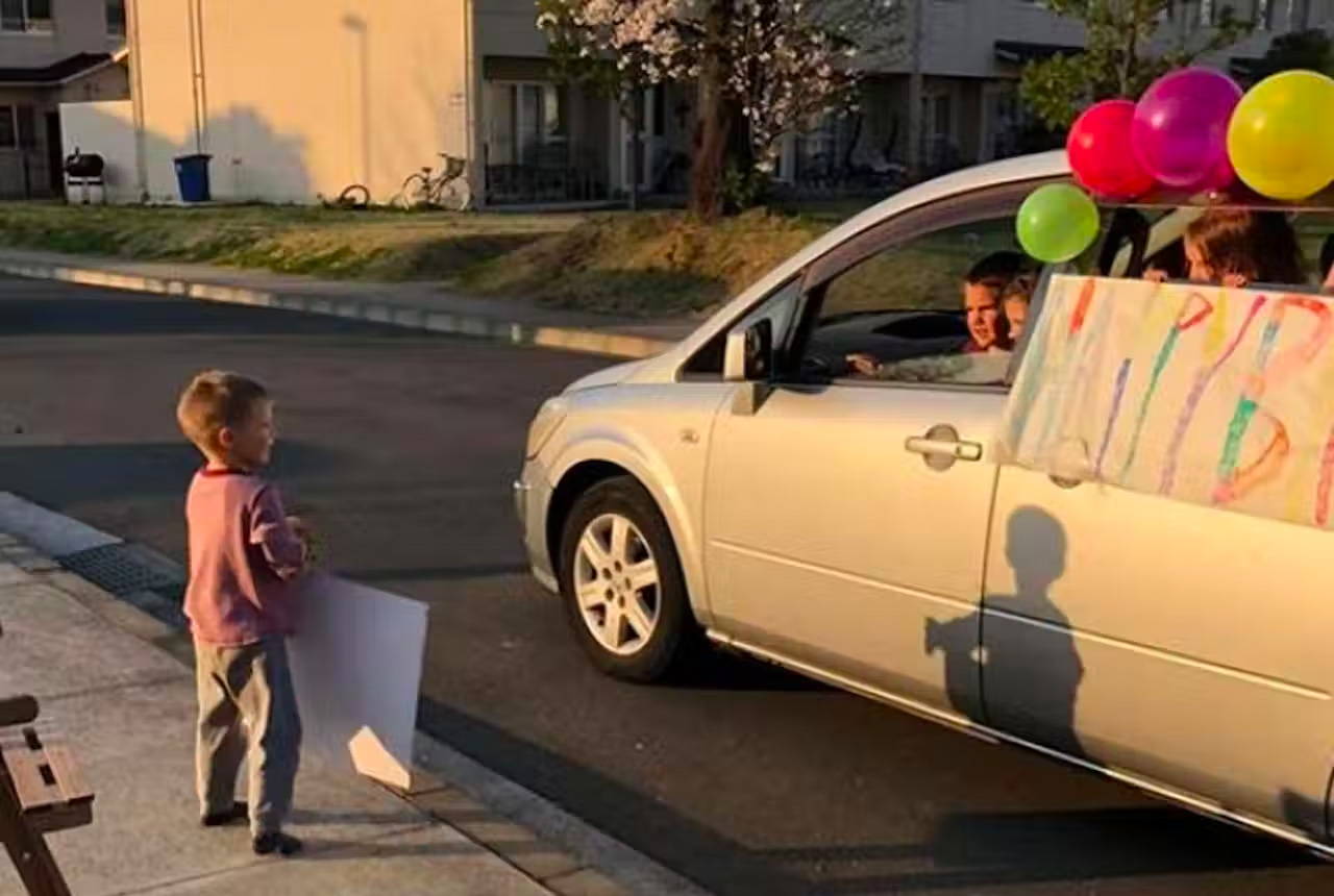 Little boy holding a poster, on a neighborhood street talking to friends driving by in a van. The van is decorated with balloons and a happy birthday sign.