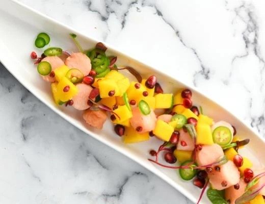 Colorful fruit salad with melon, mango, pomegranate, and green vegetables on a white plate.
