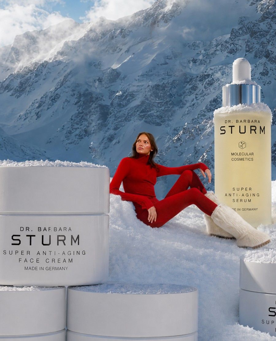 Model in red outfit beside skincare products in snowy landscape.