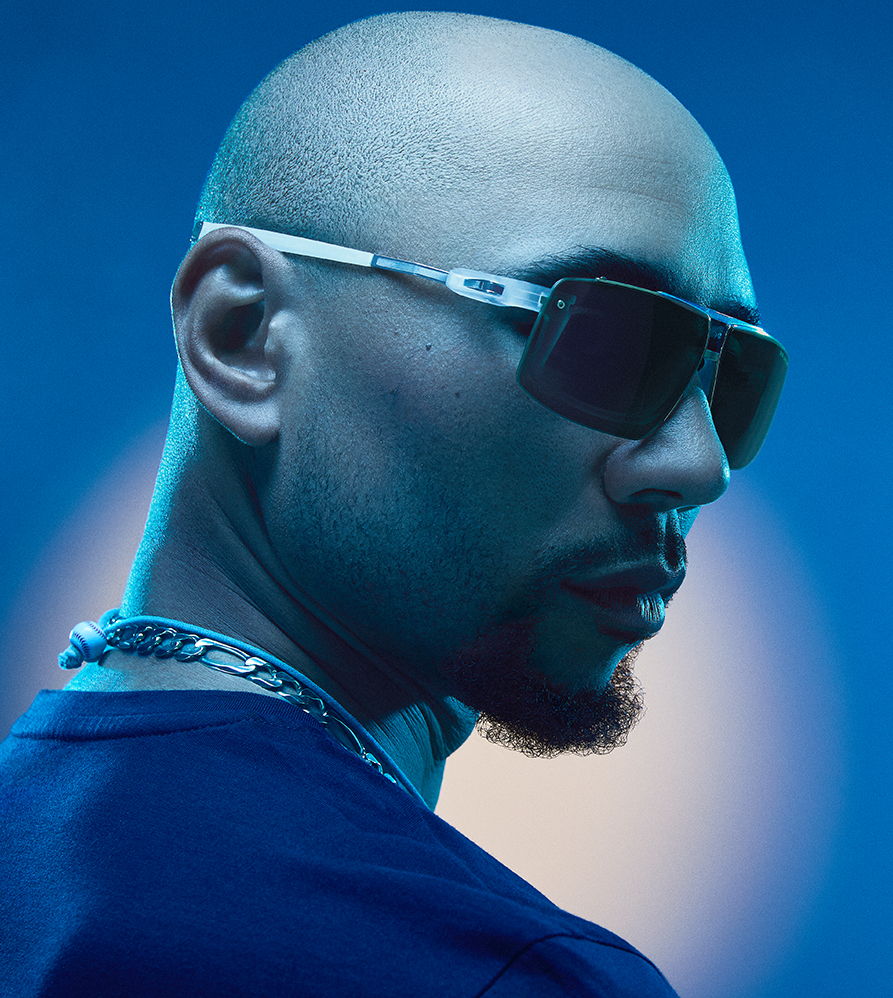 A man in sunglasses with a bald head and a chain, set against a blue background.