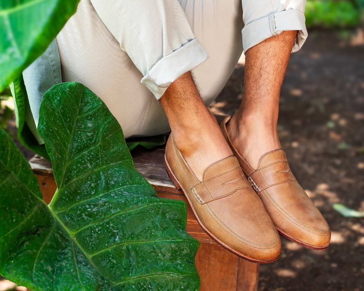 A close-up shot of a man's feet wearing tan leather Adelante Lorenzo loafers, featuring light brown distressed leather with a visible penny strap and side stitching. The man is sitting outdoors on a wooden bench or log, wearing light-colored, rolled-up chinos. A large green tropical elephant ear plant leaf is prominently featured in the lower-left foreground, and the background is a naturally lit, slightly blurred outdoor garden setting with green foliage.