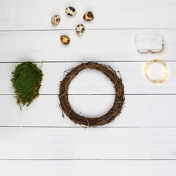 Wreath frame, foliage, decorative eggs and fairy lights against a white wooden background.