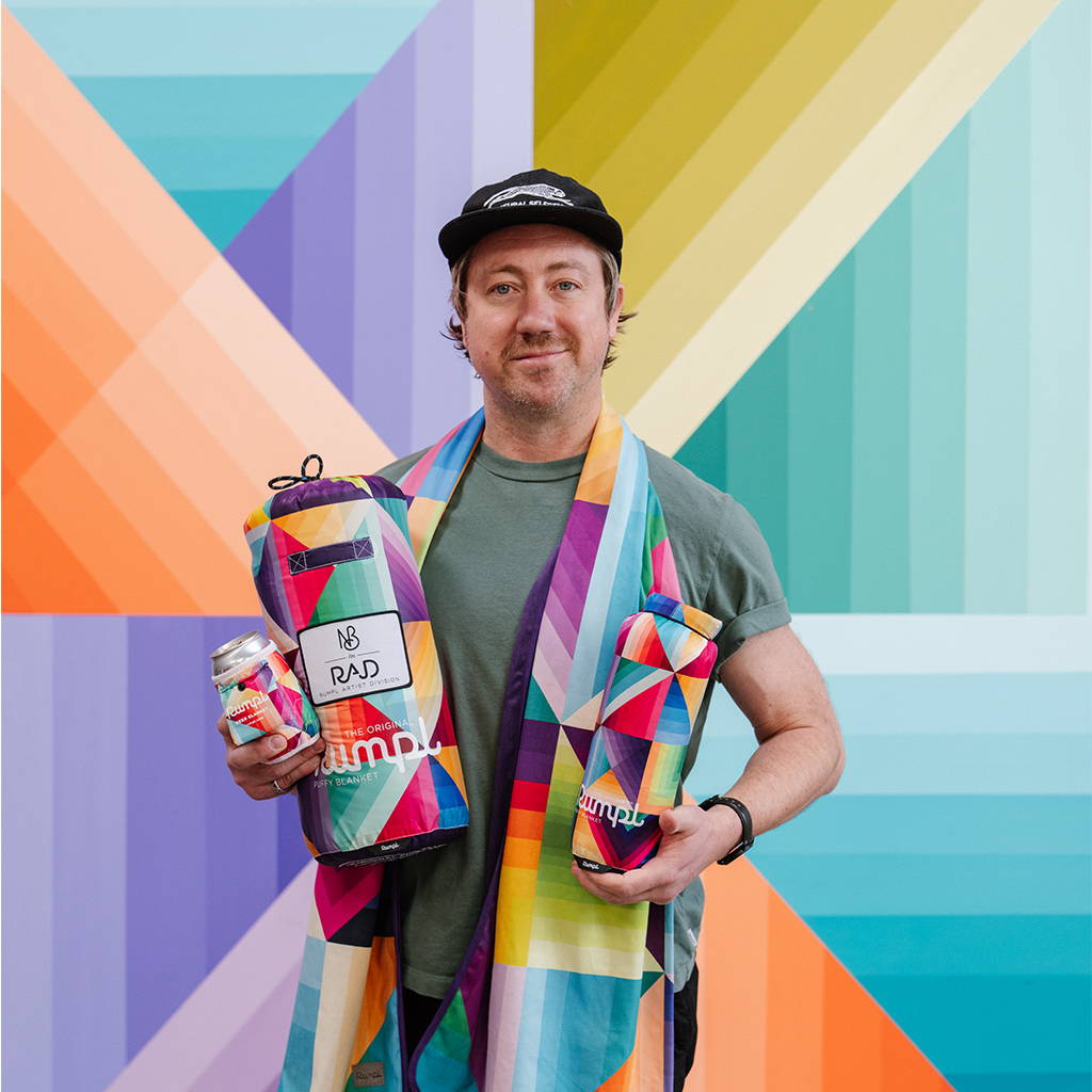 RAD Artist Nathan Brown poses in front of his mural at the Rumpl HQ while holding the Cozy Dimensions collection items.