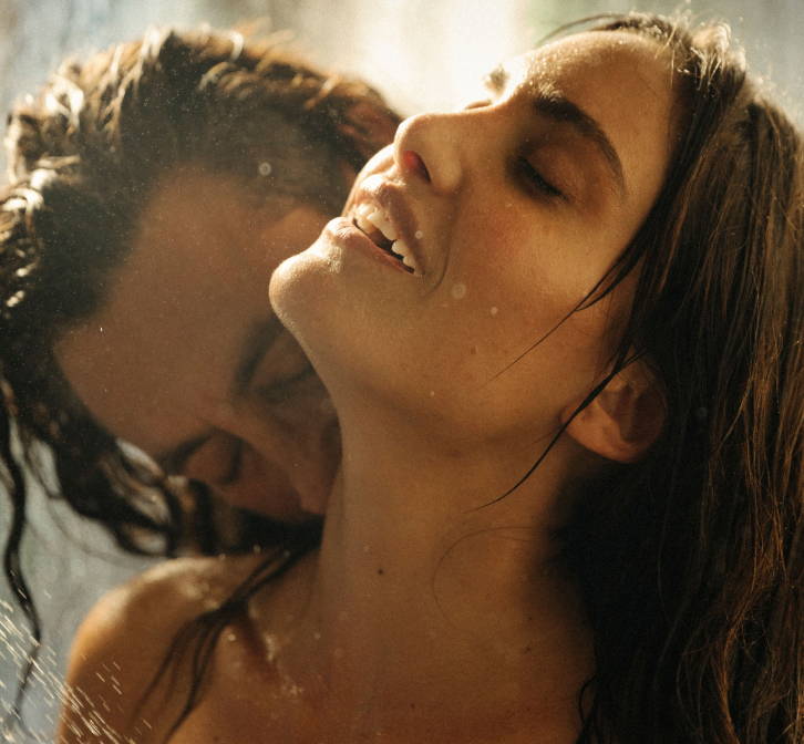 A close-up intimate moment showing a couple in the shower, with water droplets cascading down, highlighting the woman's serene expression as she tilts her head back and the man kisses her neck.
