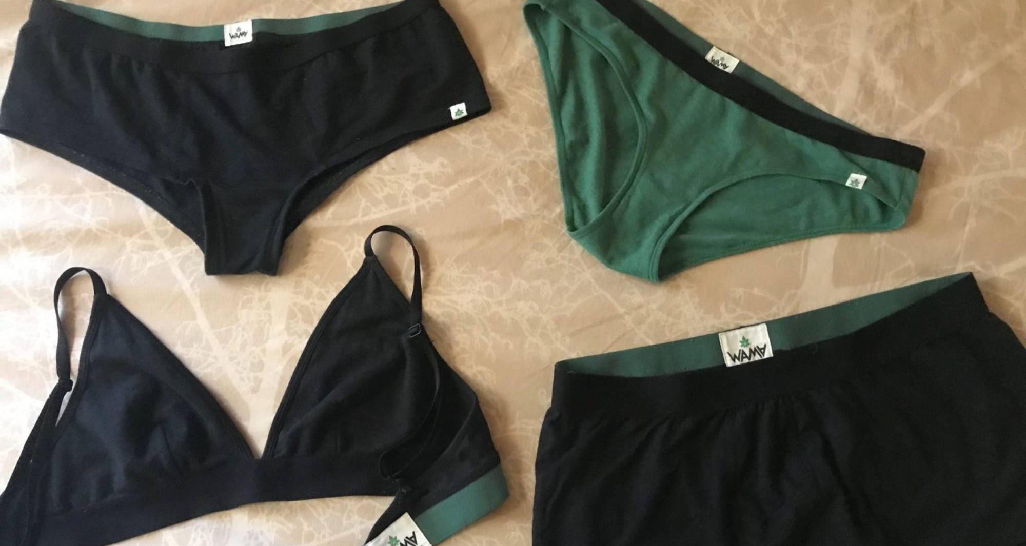 A black bralette, green underwear, black underwear, and black boy shorts laid out on a cream and white patterned sheet. 