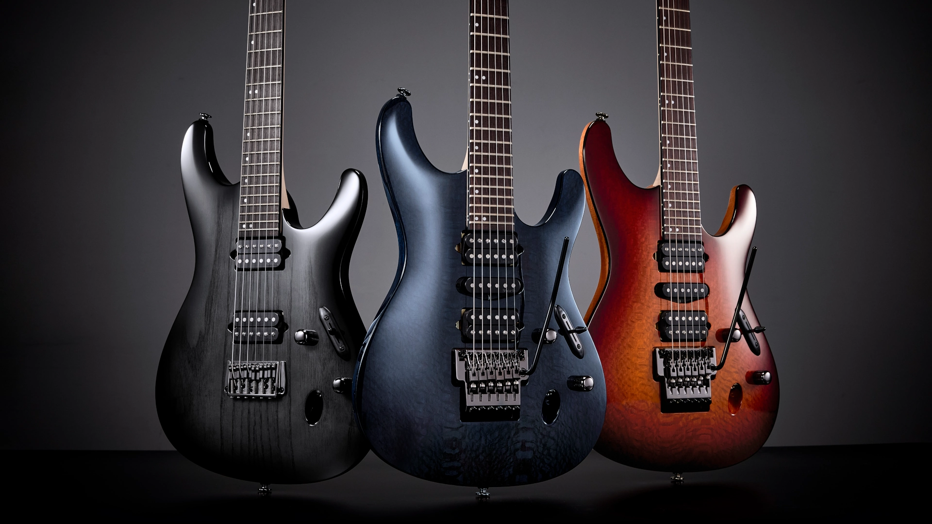 Three Ibanez S series guitars in various colours against a dark background.