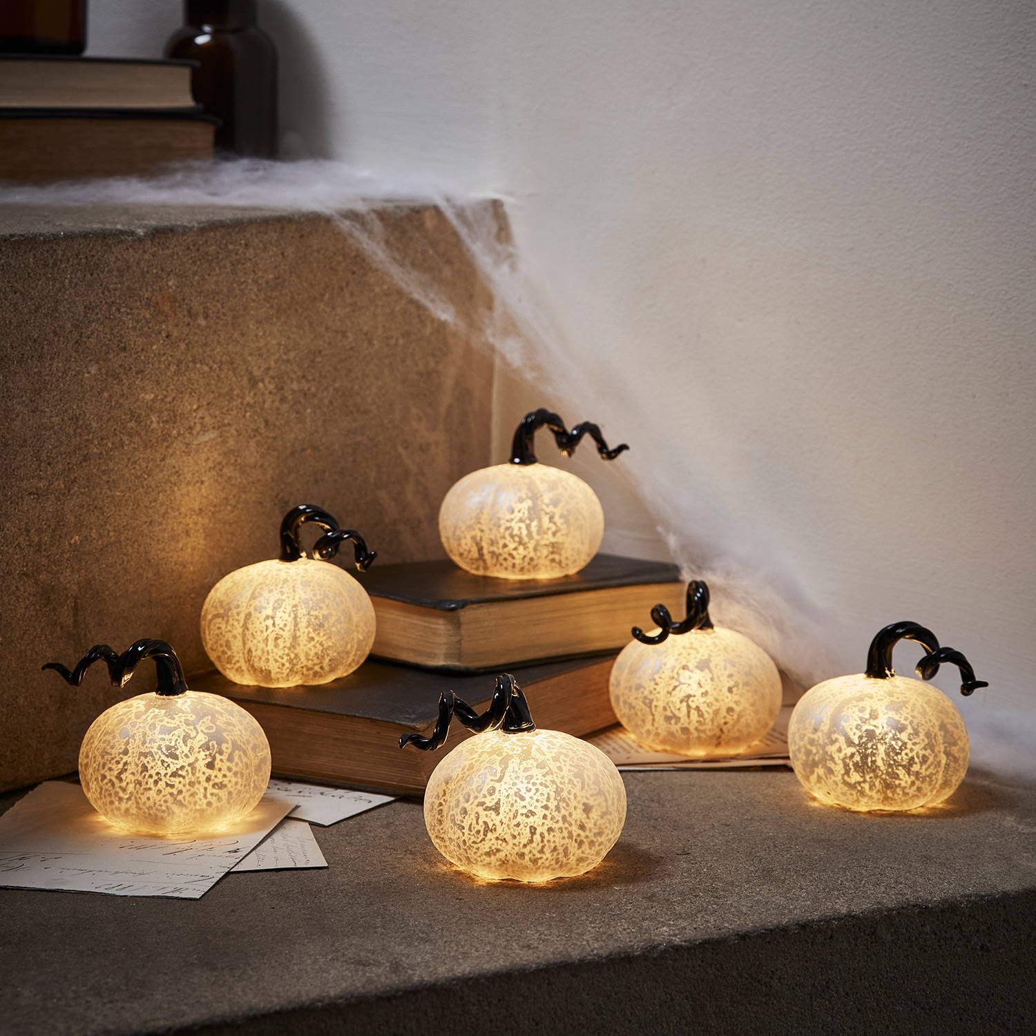 6 mini white mottled pumpkins with a black spiralled stalk on a staircase with cobwebs and books behind them.