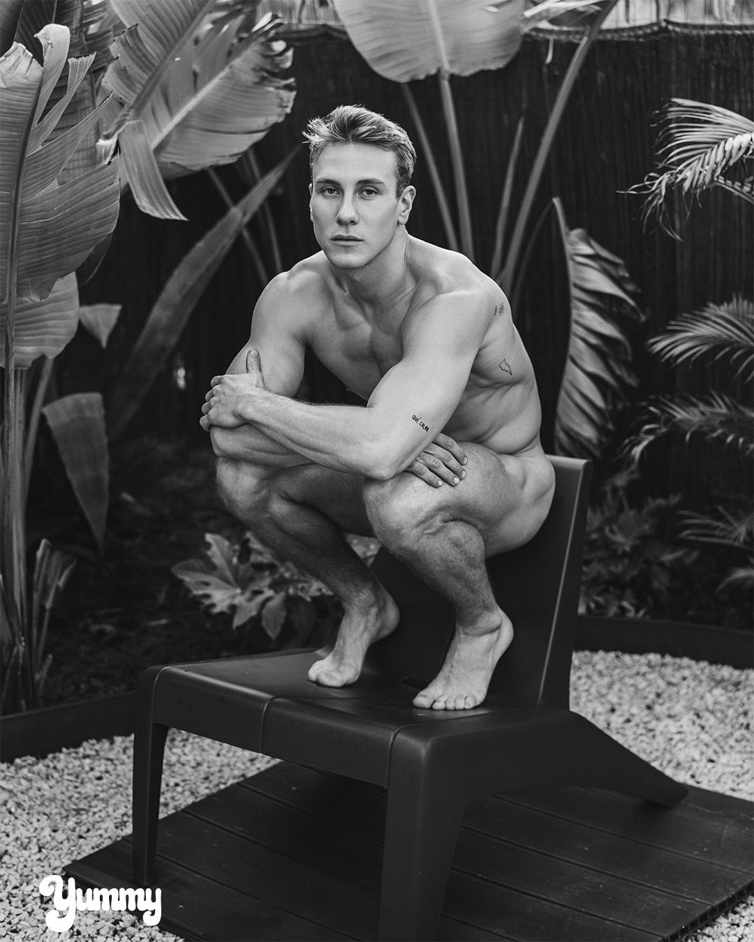 A person squatting on a chair surrounded by tropical plants in black and white.