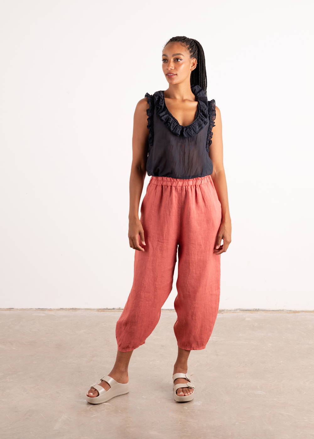 A model wearing a pair of dark pink linen barrel leg cropped trousers with a dark blue frilled sleeveless top and chunk off white platform slides