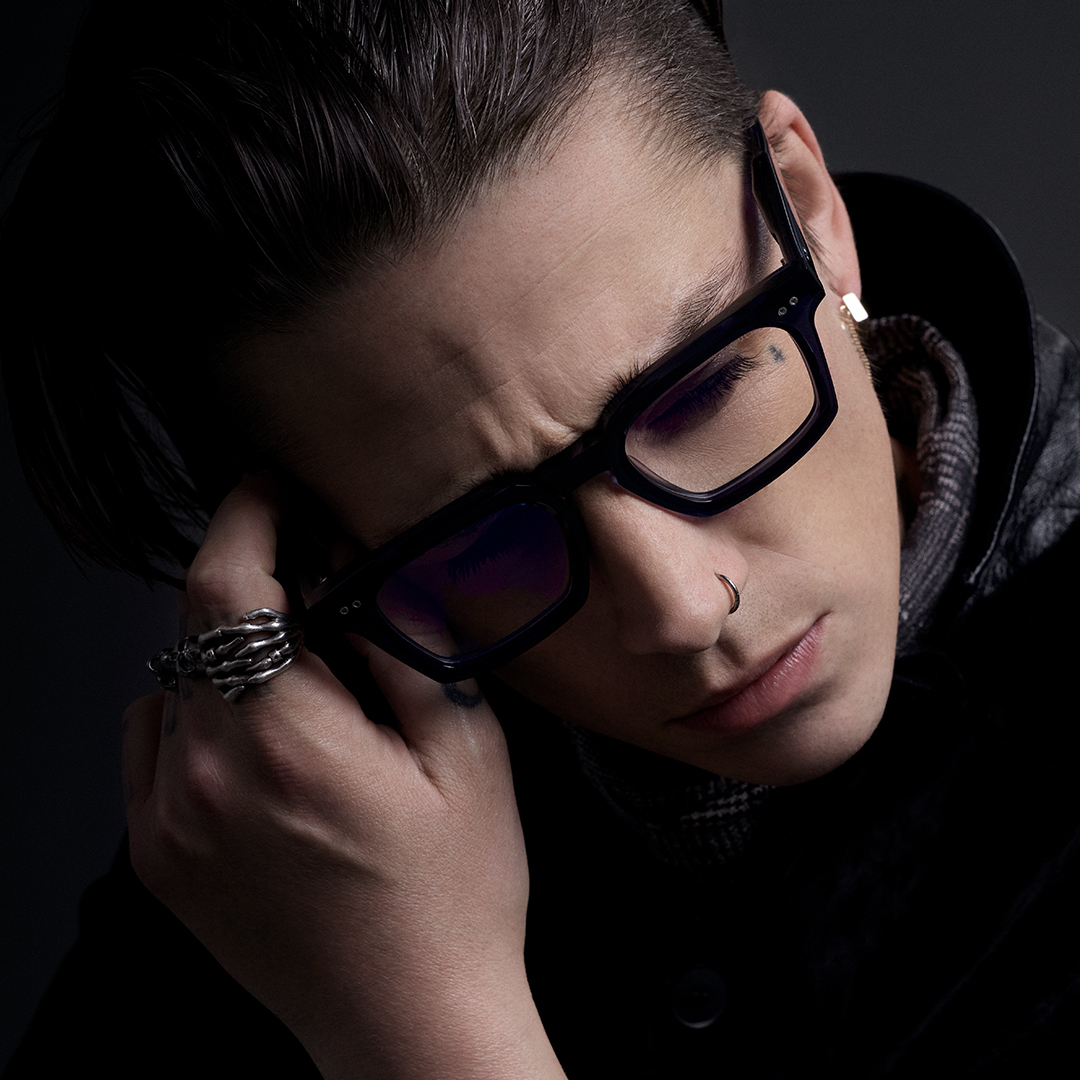 Profile of a person wearing sunglasses and a tattoo, against a dark background.