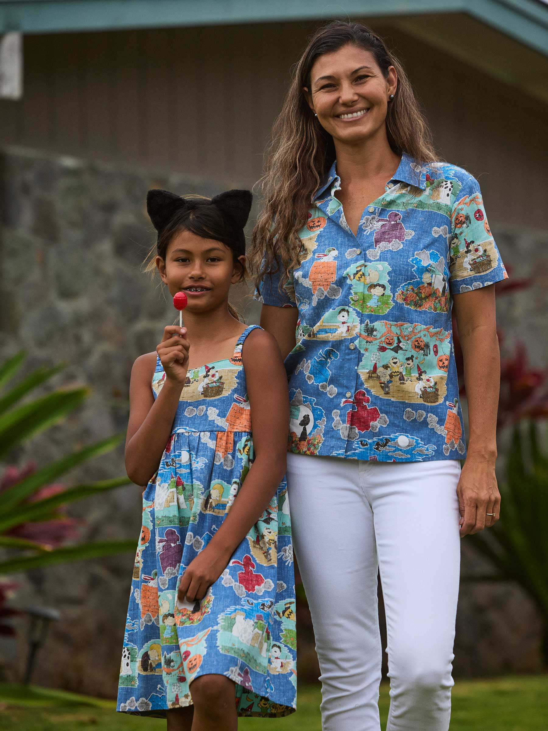 Woman and girl in matching colorful outfits, standing outdoors with a lollipop.