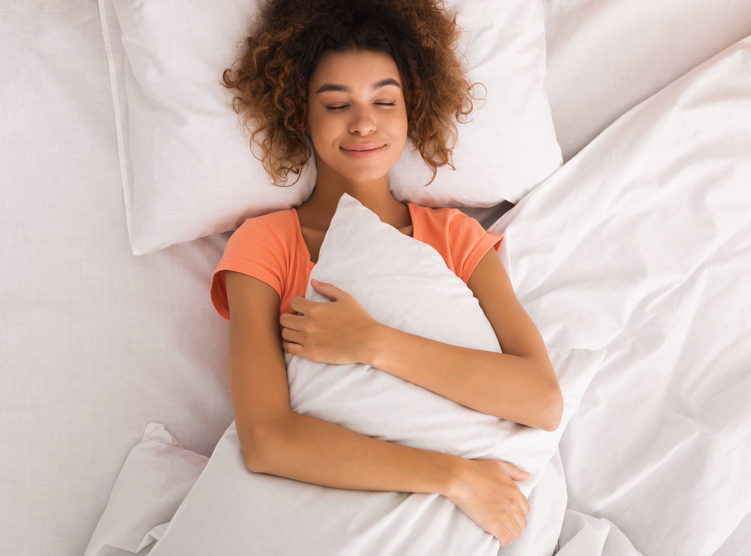 A women in an orange shirt laying down in bed hugging a copper-infused pillow while smiling.