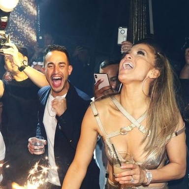 A woman in a shimmering dress lights a cake surrounded by party guests.