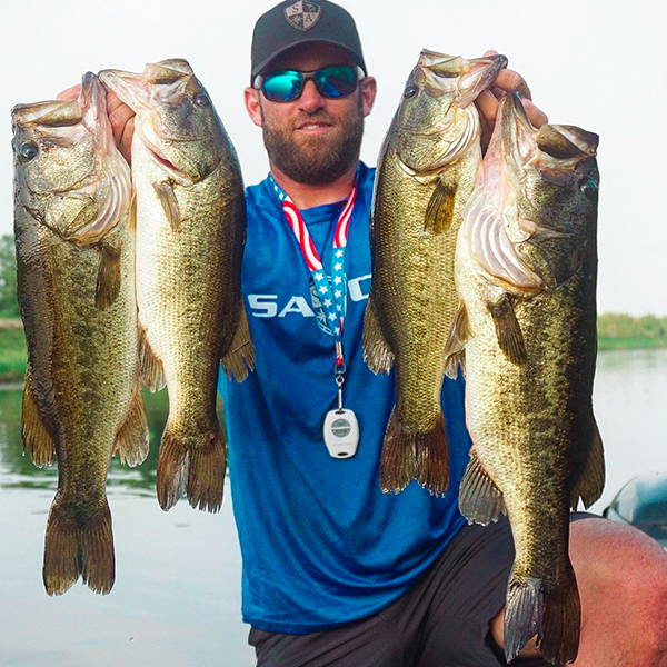 Ryan Copenhaver holding 4 bass fish, two in each hand, to the camera while wearing an SA Company performance shirt and hat.