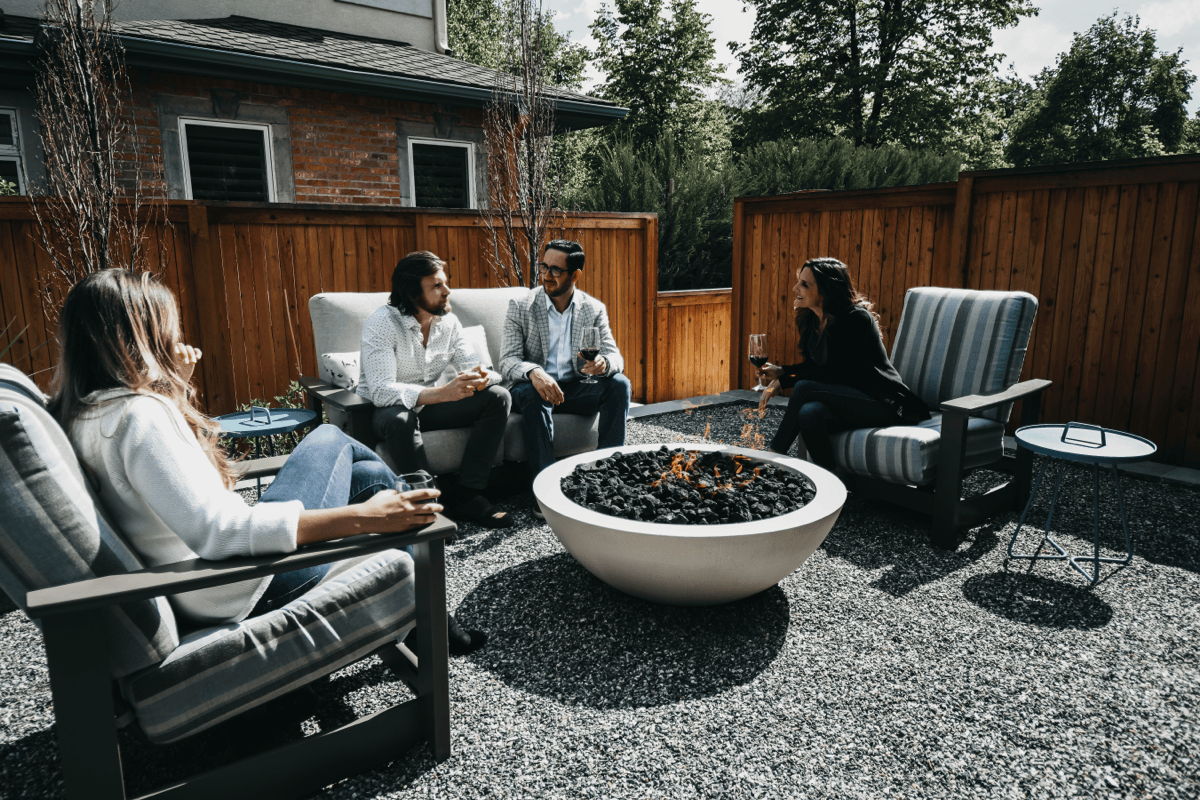 Four friends sitting in cushioned outdoor lounge chairs around a round concrete gas fire pit on a gravel patio with side tables.