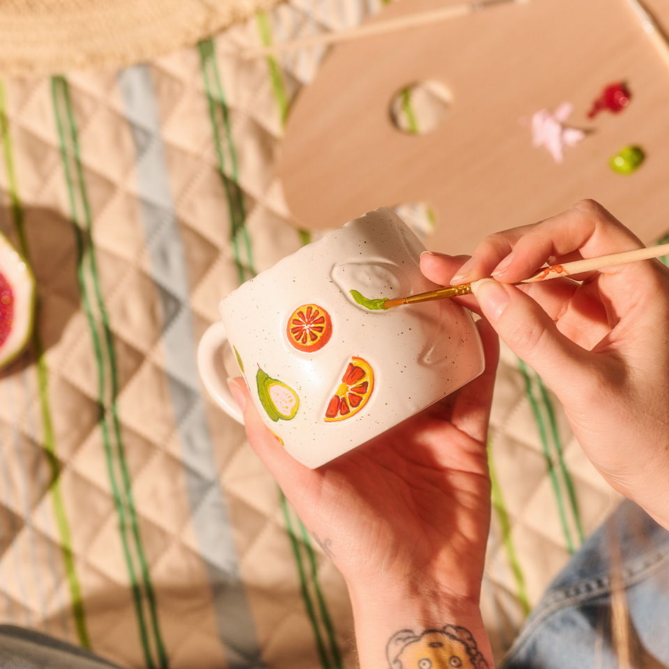 Person painting a white mug with colorful fruit designs.
