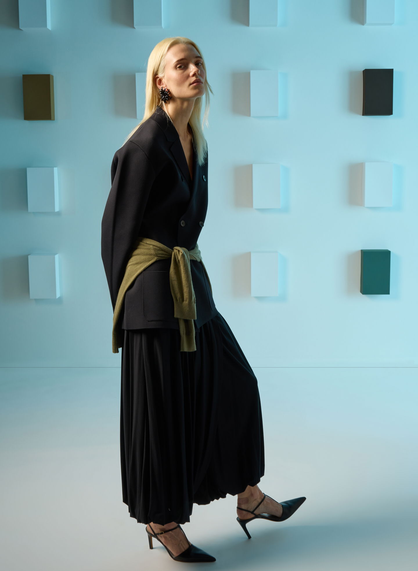 Model in black outfit with green sash, posing against abstract background.