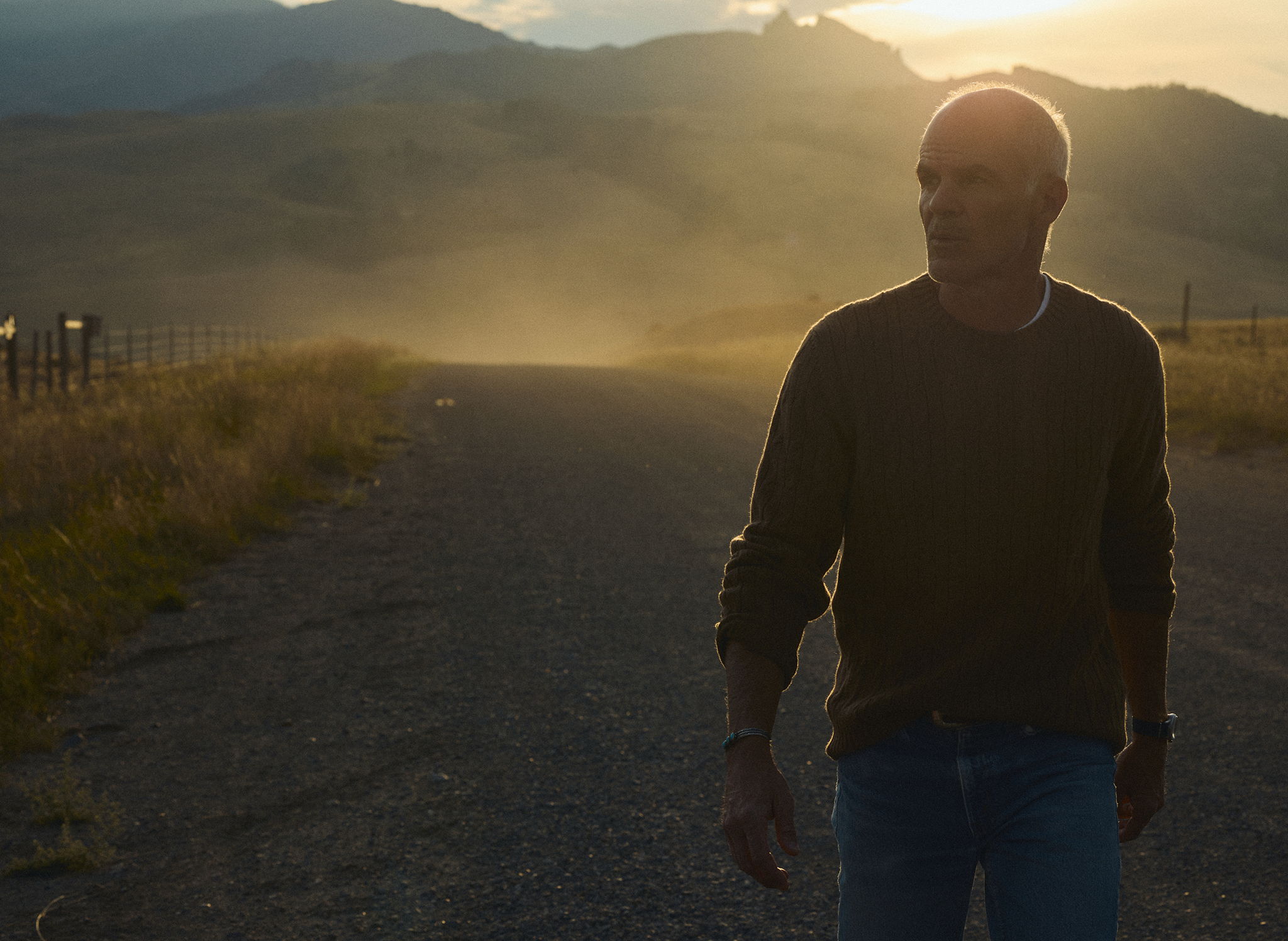 Michael Kelly in The Rivay Bowen Cashmere Cable Sweater in Saddle Brown walking down a dusty road at sunset