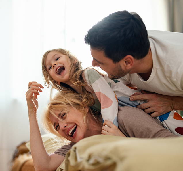 Ein kleines Mädchen spielt und kichert mit ihren Eltern beim Einschlafritual. Das feuchtwarme Klima in Betten ist ideal für Hausstaubmilben, deren Absonderungen ein häufiger Allergieauslöser sind.