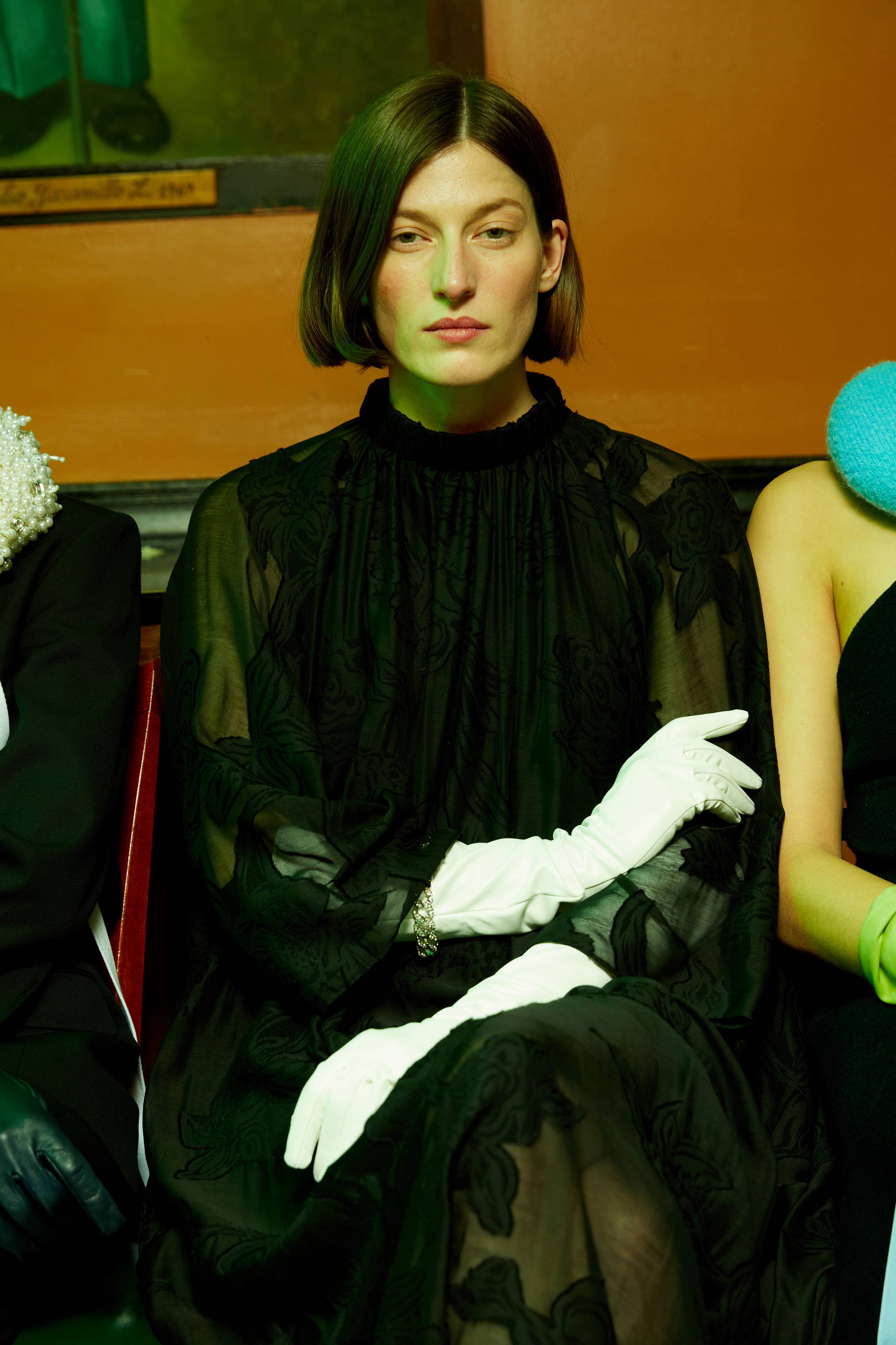 A woman in a black dress and white gloves sits calmly.